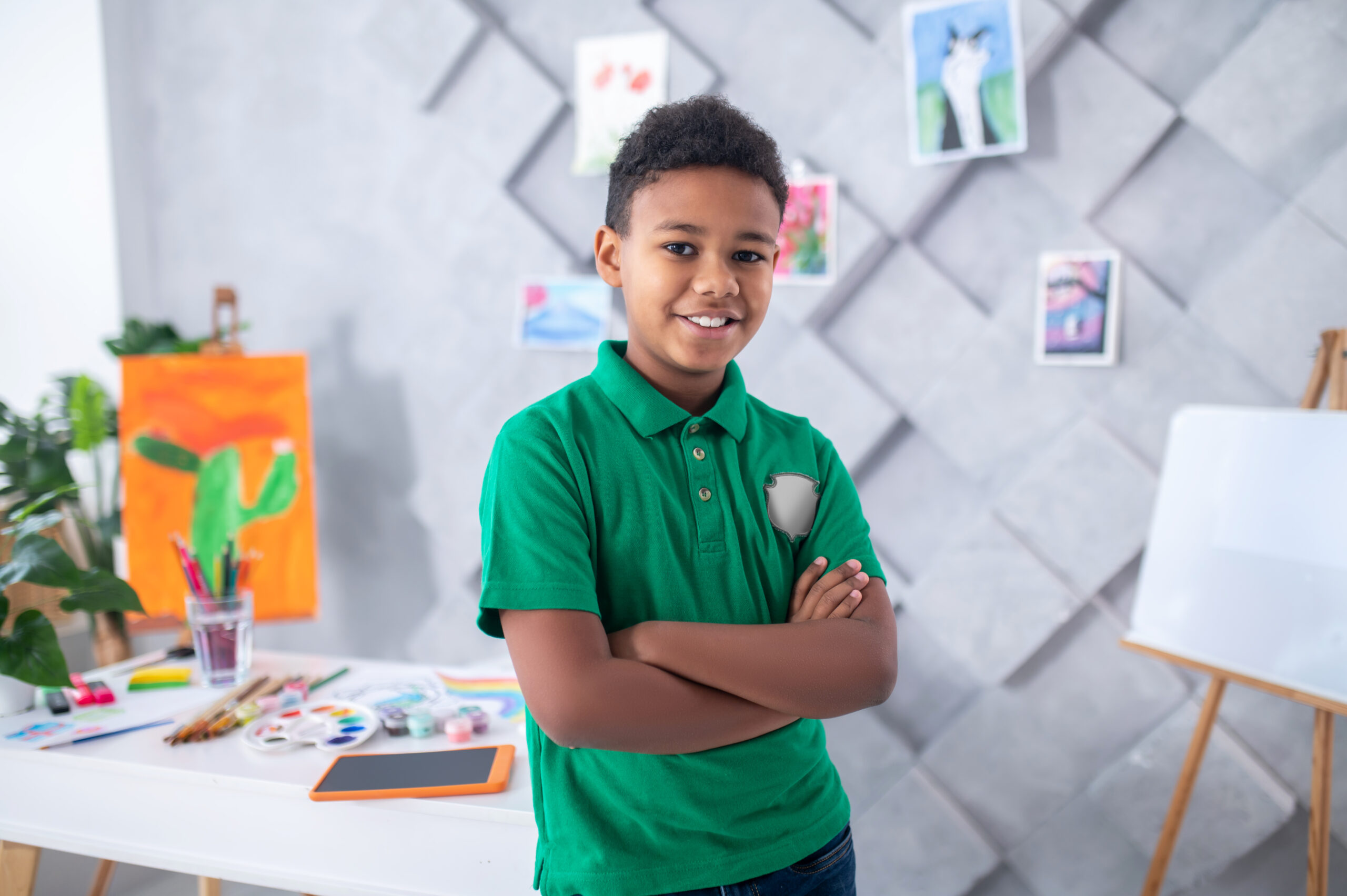 Confidence. Smiling african american boy of primary school age standing with folded arms looking at camera in art supplies room during daytime